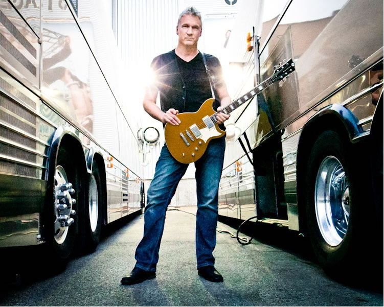 A man holding a guitar in front of two buses.